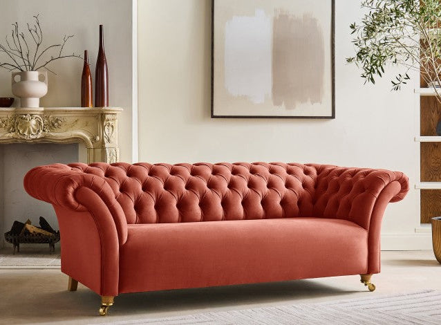 A pink Chesterfield sofa with a tufted backrest and gold-colored metal legs placed in a room with a light-colored wall and wooden flooring.