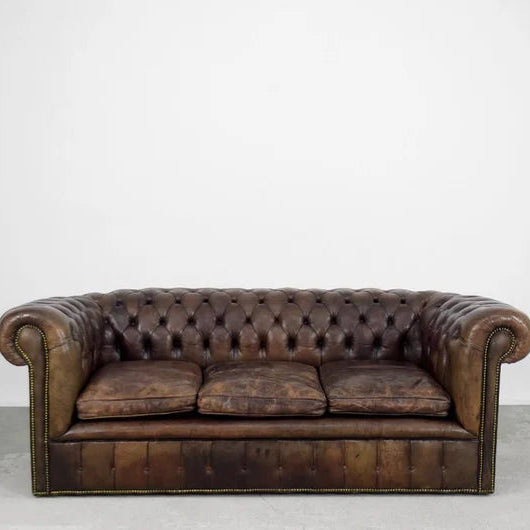 A brown three-seater Chesterfield tufted sofa made of leatherette with a wooden frame, polished teak wood legs, and beige cushions.