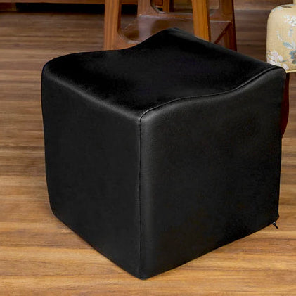 Black cube-shaped ottoman on a wooden floor with a floral-patterned chair and wooden shelf in the background.